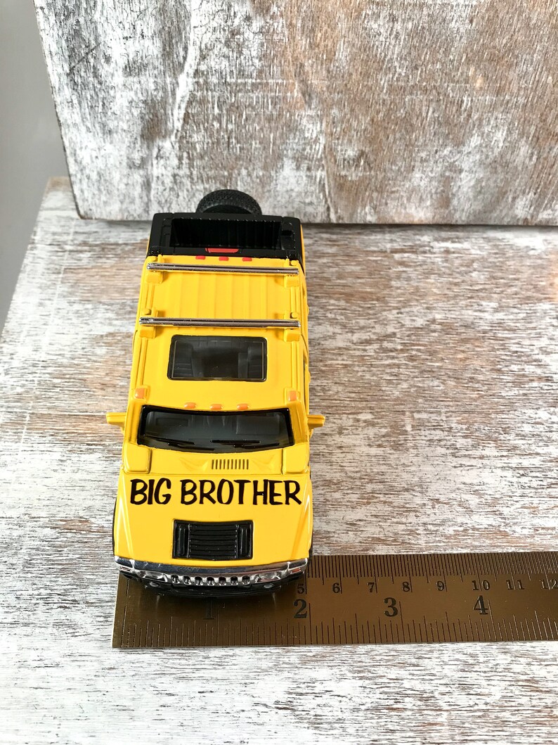 May include: Yellow toy Hummer with black details and the words "BIG BROTHER" on the hood. The toy car is sitting on a wooden surface with a ruler next to it.