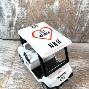 May include: White golf cart ornament with a red heart that says "Just Married" and the initials "N&H" on top. The cart has a black steering wheel and black trim. The ornament has a 2023 date on the top.