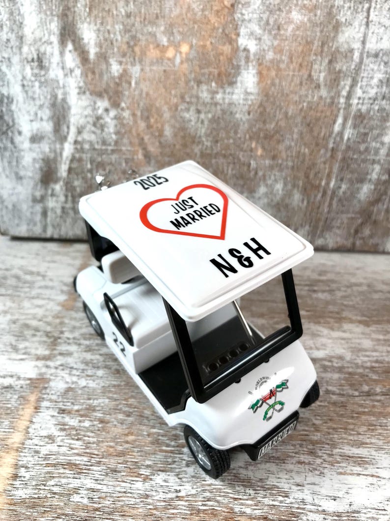 May include: White golf cart ornament with a red heart that says "Just Married" and the initials "N&H" on top. The cart has a black steering wheel and black trim. The ornament has a 2023 date on the top.