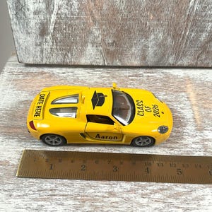 Puede incluir: Un coche de juguete amarillo con el texto "CLASS OF 2026" y "Aaron" impreso. El coche tiene un gorro de graduaci&oacute;n negro en la parte superior y las palabras "DATE HERE" en la parte trasera. Una regla est&aacute; al lado del coche.