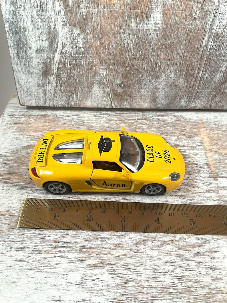 Puede incluir: Un coche de juguete amarillo con el texto "CLASS OF 2026" y "Aaron" impreso. El coche tiene un gorro de graduaci&oacute;n negro en la parte superior y las palabras "DATE HERE" en la parte trasera. Una regla est&aacute; al lado del coche.