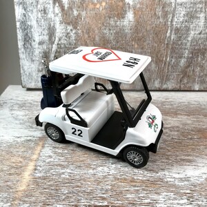 May include: White golf cart toy with a red heart and the words "Just Married" on the roof. The cart has a black steering wheel, black seats, and black wheels. The cart has a golf bag hanging on the side and the number 22 on the side.