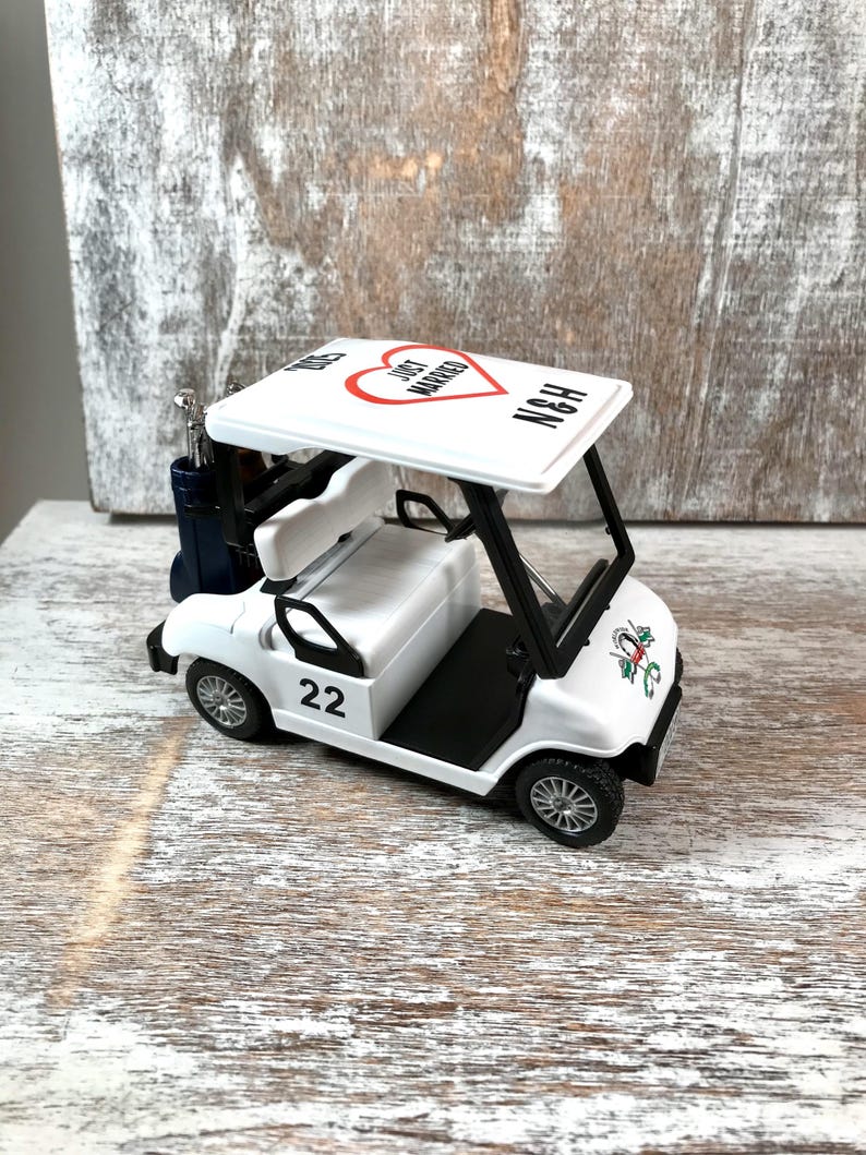 May include: White golf cart toy with a red heart and the words "Just Married" on the roof. The cart has a black steering wheel, black seats, and black wheels. The cart has a golf bag hanging on the side and the number 22 on the side.