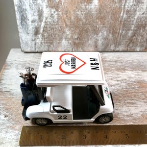 May include: White golf cart toy with a red heart that says "Just Married" on the roof. The cart has a golf bag with clubs and the number 22 on the side. The roof also says "N&H" and "2023".
