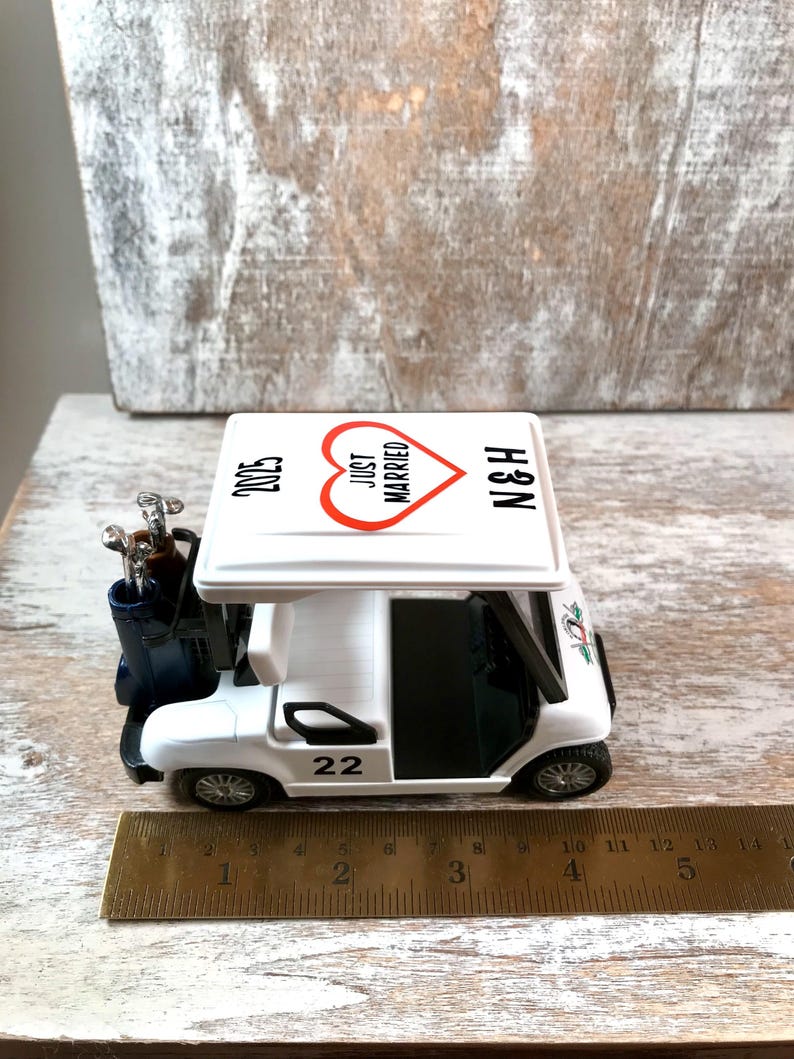 May include: White golf cart toy with a red heart that says "Just Married" on the roof. The cart has a golf bag with clubs and the number 22 on the side. The roof also says "N&H" and "2023".