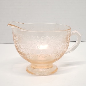 May include: A vintage pink glass creamer with a delicate floral pattern. The creamer has a footed base and a small handle.