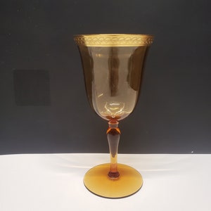 Peut inclure: Verre à vin ambré avec un bord et un pied dorés. Le verre a une large coupe et une tige fine, et la base est un cercle plat. Le bord présente un motif doré décoratif. Le verre est posé sur un fond noir et blanc.