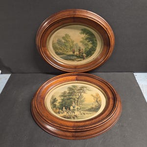 May include: Two oval wall frames made of polished brown wood, each containing a vintage-style landscape print. The top print depicts figures near a river, while the bottom print shows people and a horse crossing a stream amidst green trees.