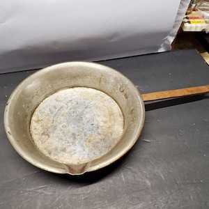 May include: A vintage copper pan with a wooden handle. The pan has a spout and is marked with numbers on the side.