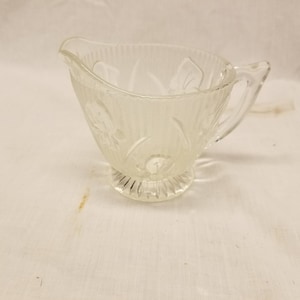 May include: A clear glass creamer with a decorative floral pattern and a handle. The creamer has a fluted base.
