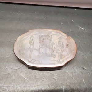 Könnte beinhalten: Eine ovale, dunkelbraune Holzkiste mit einem gravierten Design von drei Figuren in einer Kutsche. Die Kiste hat einen leicht erhöhten Rand und eine glatte Oberfläche.