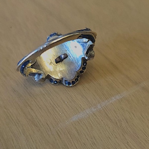 May include: A gold-toned ring with a decorative design. The ring features a central setting with a small, rectangular element and is adorned with small, dark blue accents around the edges. The ring is set against a light brown wooden surface.