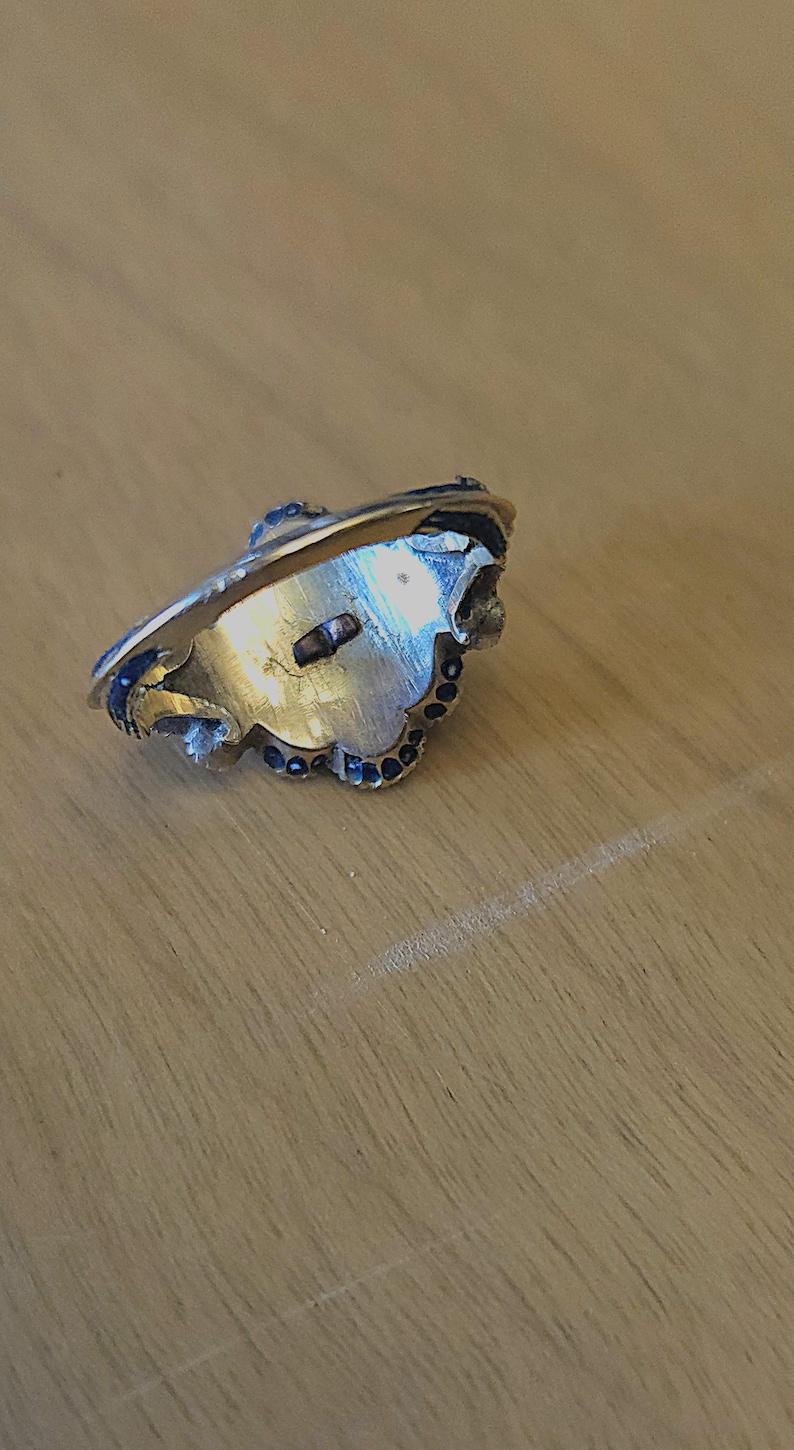 May include: A gold-toned ring with a decorative design. The ring features a central setting with a small, rectangular element and is adorned with small, dark blue accents around the edges. The ring is set against a light brown wooden surface.