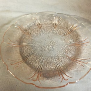 May include: A clear, pink glass plate with a floral pattern. The plate has a scalloped edge and a detailed embossed design featuring flowers and leaves. The plate is likely vintage or antique, and is suitable for serving food.