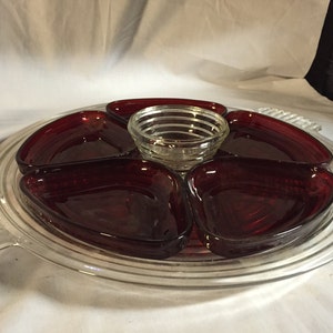 May include: A clear glass serving tray with a raised rim and two handles. The tray has five red glass bowls in the shape of hearts. The bowls are arranged in a circle around a smaller clear glass bowl in the center.