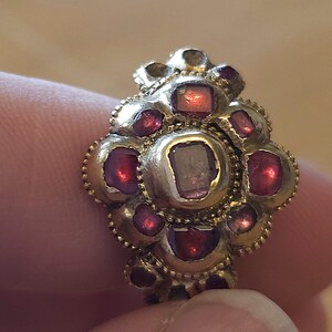 May include: A gold-toned ring with a floral design, featuring a central stone and surrounding red gemstones. The ring's intricate details and the contrast of colors create a striking visual appeal. A piece of jewelry.