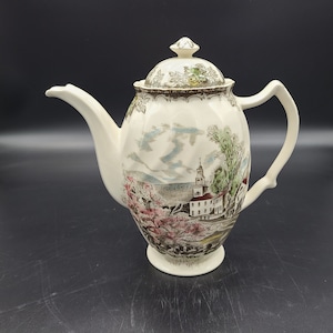 Puede incluir: Tetera de cerámica color crema con tapa, asa y boquilla. La tetera presenta un diseño de paisaje escénico con una iglesia, árboles y montañas en tonos verdes, rosas y azules. La tapa está rematada con un adorno decorativo.