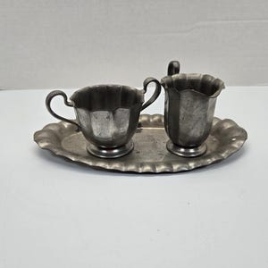 Könnte beinhalten: Ein silbernes Sahnegießer- und Zuckergefäß-Set mit passendem Tablett. Das Set hat ein gewelltes Design und ist aus Zinn.