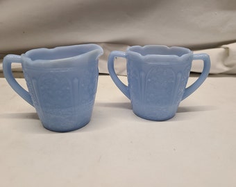 May include: A light blue glass creamer and sugar bowl set. The pieces have a floral pattern and scalloped edges. The creamer has a handle and a spout. The sugar bowl has two handles.