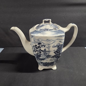 Puede incluir: Tetera de cerámica blanca con un diseño de paisaje azul y blanco. La tetera tiene un pico curvo, un asa y una tapa con un pequeño asa. El diseño presenta montañas, árboles y edificios.