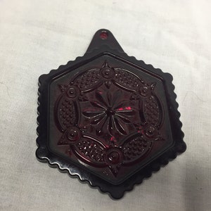 May include: A dark red, hexagonal decorative piece with a scalloped edge and a hole for hanging. The centre features a starburst design surrounded by floral and dot patterns. The piece is made of a translucent material.