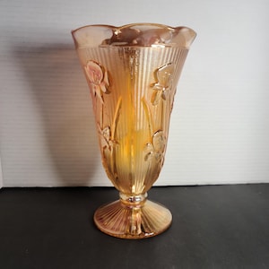 May include: An iridescent amber glass vase with a flared top and a pedestal base. The vase features embossed floral designs and vertical ribbed detailing. The glass has a shimmering, opalescent quality, reflecting light with a warm, golden hue. A decorative piece.
