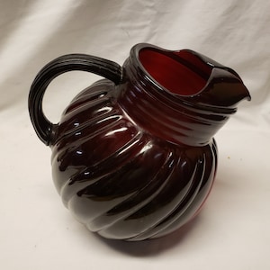 May include: A dark red glass pitcher with a black plastic handle. The pitcher has a ribbed design and a wide mouth.