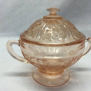 May include: A pink glass sugar bowl with a lid, featuring a floral design. The bowl has two handles and sits on a pedestal base.