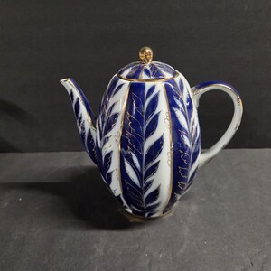 May include: A porcelain teapot featuring a white base with vertical panels of deep blue leaf patterns outlined in gold. The teapot has a curved handle, a pointed spout, and a matching lid topped with a small gold-colored spherical knob.