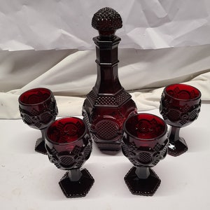 May include: A ruby red glass decanter with a matching stopper and four goblets. The set features a textured, geometric design. The decanter is tall and slender, while the goblets have a hexagonal base.