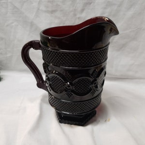 May include: A ruby-red glass pitcher with a decorative pattern. The pitcher features a handle and a spout for pouring. The design includes diamond and floral patterns. A vintage-style piece for serving beverages.