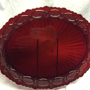 May include: A ruby red, oval-shaped glass serving platter. The platter has a scalloped edge with a repeating pattern of oval and dot designs. The center of the platter has a sunburst design.