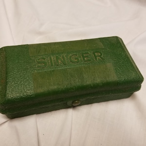 Könnte beinhalten: Eine grüne rechteckige Box mit dem Wort "SINGER" in Prägung auf der Oberseite.