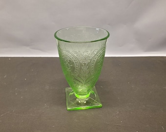 May include: A green glass goblet with a square base. The goblet has a flared rim and is decorated with an embossed floral pattern. The glass has a slight green tint and is likely vintage.