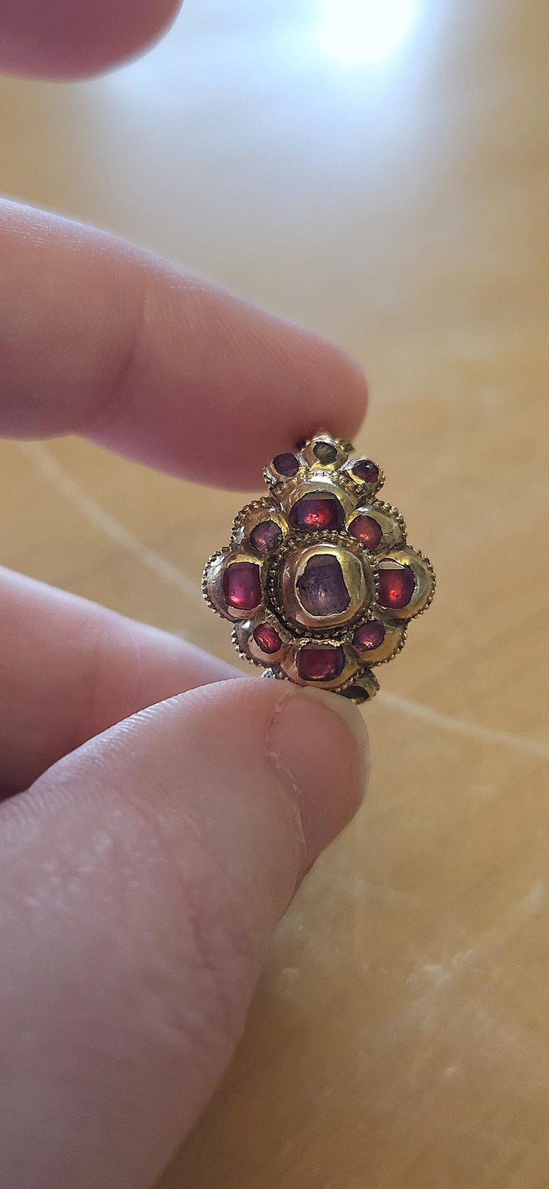 May include: A gold-toned, floral-shaped ring with multiple red gemstones. The ring's design features a central, larger gemstone surrounded by smaller stones, creating a flower-like pattern. The ring is held between fingers.