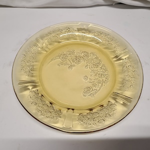 May include: A yellow glass plate with a floral design. The plate has a raised rim and a scalloped edge. The floral design is of roses and leaves.