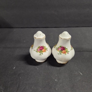 May include: Two white ceramic salt and pepper shakers with a floral rose design. The shakers have a bulbous shape with a gold rim and a small hole pattern on top. The floral design includes pink, red, and yellow roses.