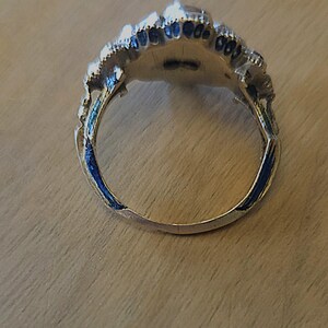 May include: A silver-toned ring with a detailed, ornate design. The ring features a central setting surrounded by a decorative border with blue enamel accents. The ring is set against a wooden surface.