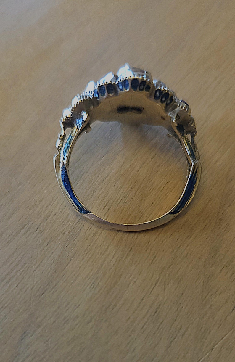 May include: A silver-toned ring with a detailed, ornate design. The ring features a central setting surrounded by a decorative border with blue enamel accents. The ring is set against a wooden surface.