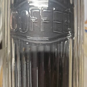 May include: A clear glass coffee canister with the word "COFFEE" embossed on the front. The canister has vertical ribbed detailing and a dark interior. The top of the canister has a decorative design.