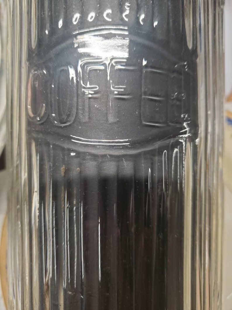 May include: A clear glass coffee canister with the word "COFFEE" embossed on the front. The canister has vertical ribbed detailing and a dark interior. The top of the canister has a decorative design.
