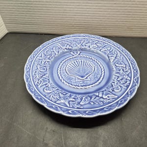 May include: A round, periwinkle blue ceramic plate featuring a central embossed scallop shell surrounded by a rope-like border. The rim is decorated with a relief pattern of various seashells, starfish, and seaweed, all in a matching blue tone.