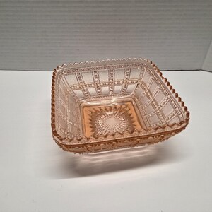 Pode incluir: Uma tigela quadrada de vidro rosa com um padrão detalhado e texturizado. A tigela tem uma borda ondulada e um design central de raio de sol. A estética geral é vintage e decorativa, adequada para servir ou exibir.