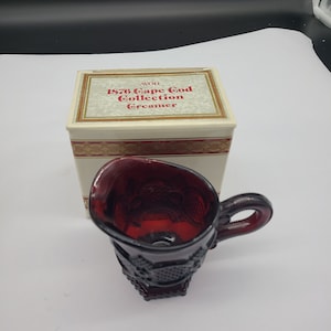 Op de afbeelding: Een rode glazen roomkan met een decoratief ontwerp. De roomkan zit in de originele doos, waarop "Avon 1876 Cape Cod Collection Creamer" staat.