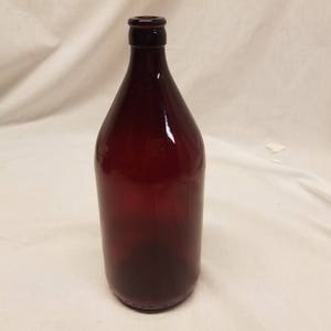 Puede incluir: Una botella alta de vidrio rojo oscuro con cuello largo y cuerpo redondeado. La botella está vacía y parece estar hecha de vidrio grueso. La botella está sobre una superficie blanca.