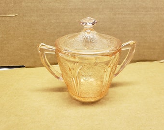 May include: A vintage, light pink glass sugar bowl with a lid. The bowl has two handles and a floral pattern etched into the glass. The lid has a faceted knob. The sugar bowl is a classic piece of glassware.
