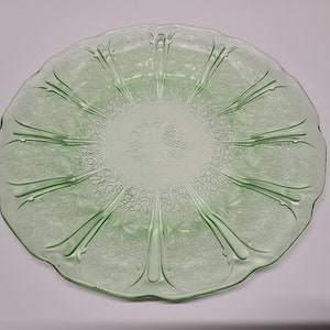 May include: A round, light green glass plate featuring a scalloped edge and a pressed floral pattern. The design includes radiating vertical ribs that divide the surface into sections, each decorated with embossed flowers and leaves.