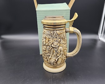 May include: A ceramic beer stein with a gold lid and handle. The stein features a detailed relief design of a man panning for gold in a forest setting.