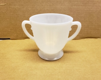 May include: A white, two-handled sugar bowl with a pedestal base. The bowl has a rounded shape with a textured pattern on the sides and handles. The sugar bowl is made of opaque white glass.