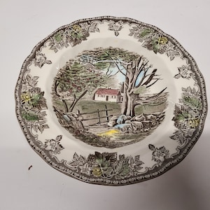 Pode incluir: Um prato de cerâmica branco com uma borda floral marrom e verde. O centro do prato apresenta uma cena de casa de campo com uma árvore, uma casa e uma cerca.